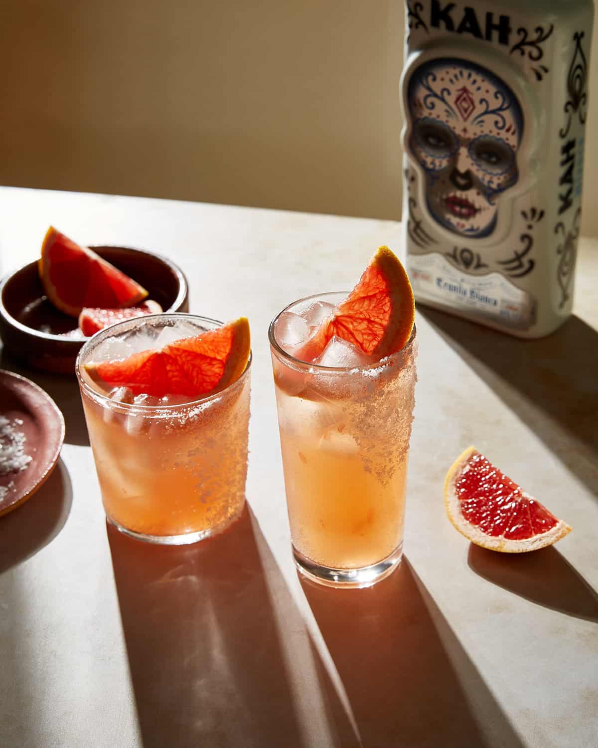 two glasses with paloma cocktail garnished with slice of grapefruit and blanco tequila bottle in background